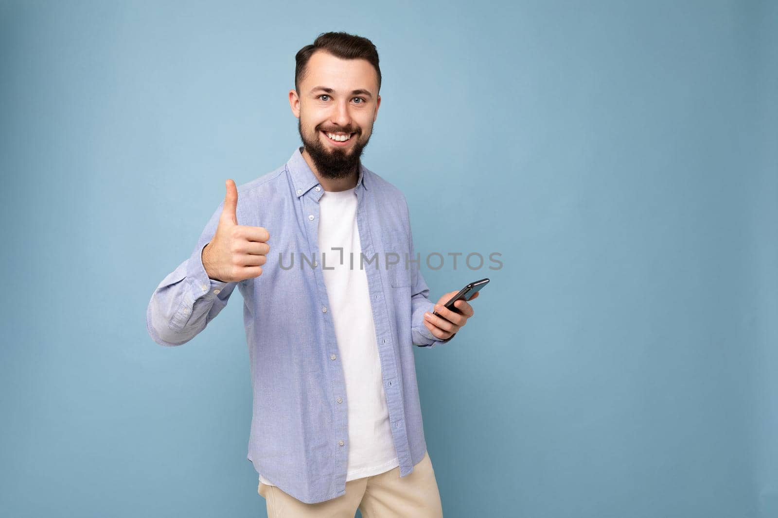 Joyful handsome young brunette unshaven man with beard wearing stylish white t-shirt and blue shirt isolated over blue background with empty space holding in hand and using phone messaging sms looking at camera giving thumbs up gesture.