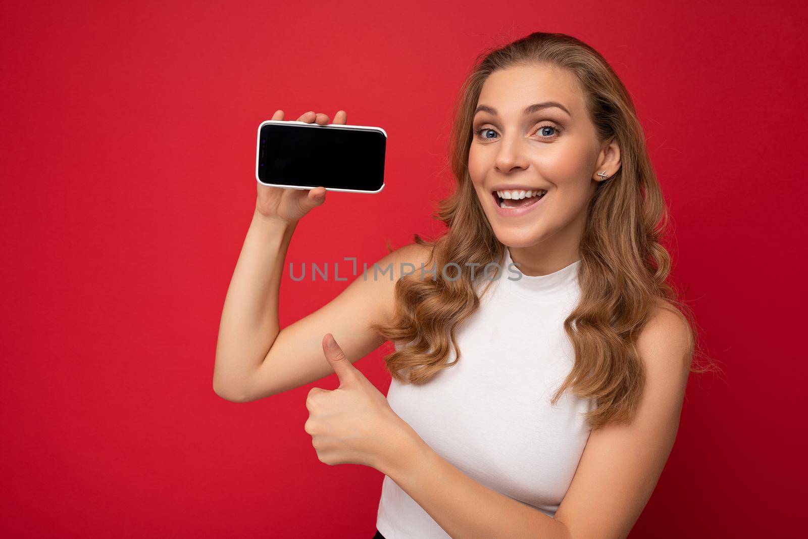 Closeup of happy smiling beautiful young blonde woman wearing white t-shirt isolated on red background with copy space holding smartphone showing phone in hand with empty screen for mockup looking at camera and showing thumbs up gesture.