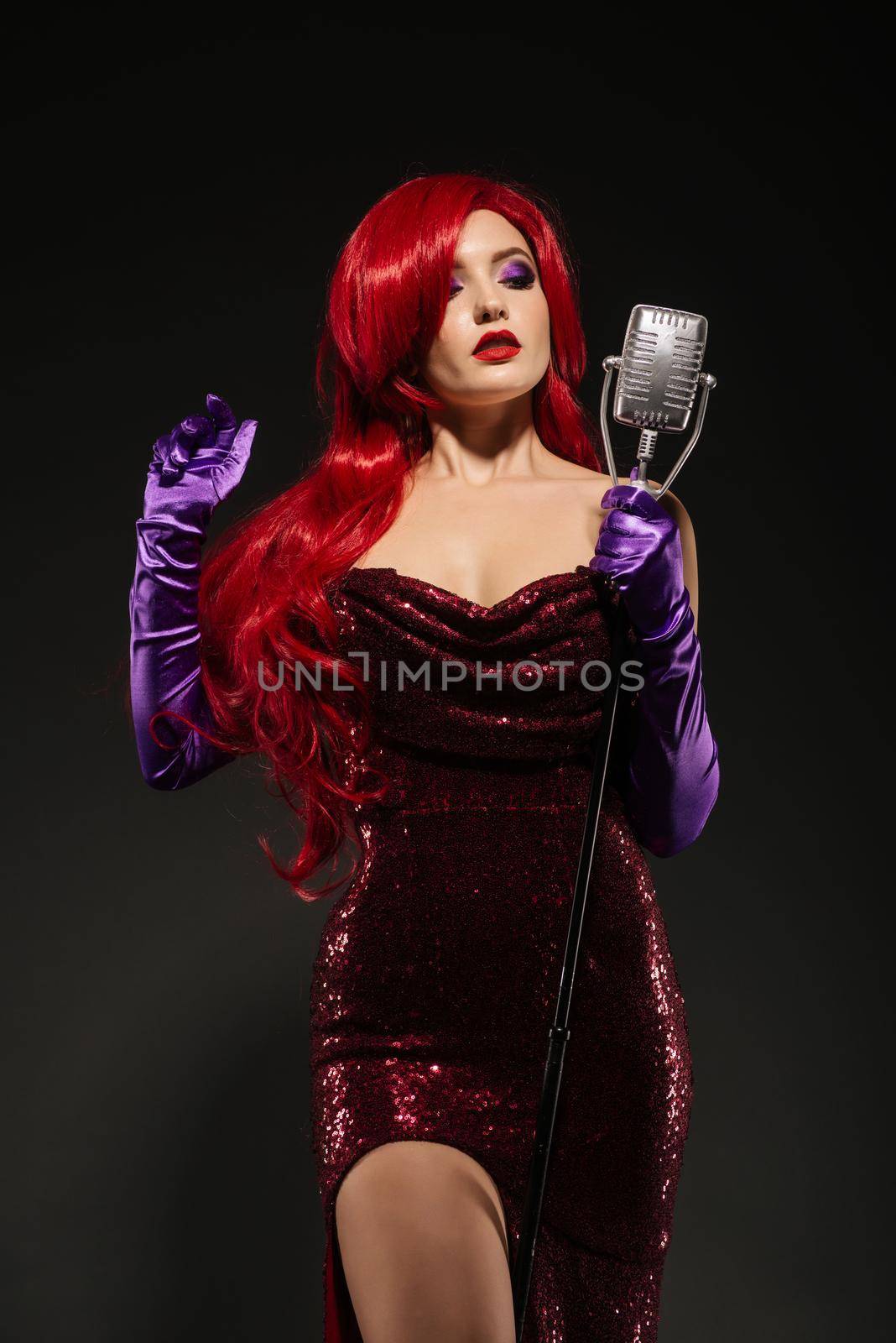 Young beauty redhead woman with very long hair in red gown with microphone on the stand on a black background. Sexy gown on a beautiful slim figure. Long pink gloves