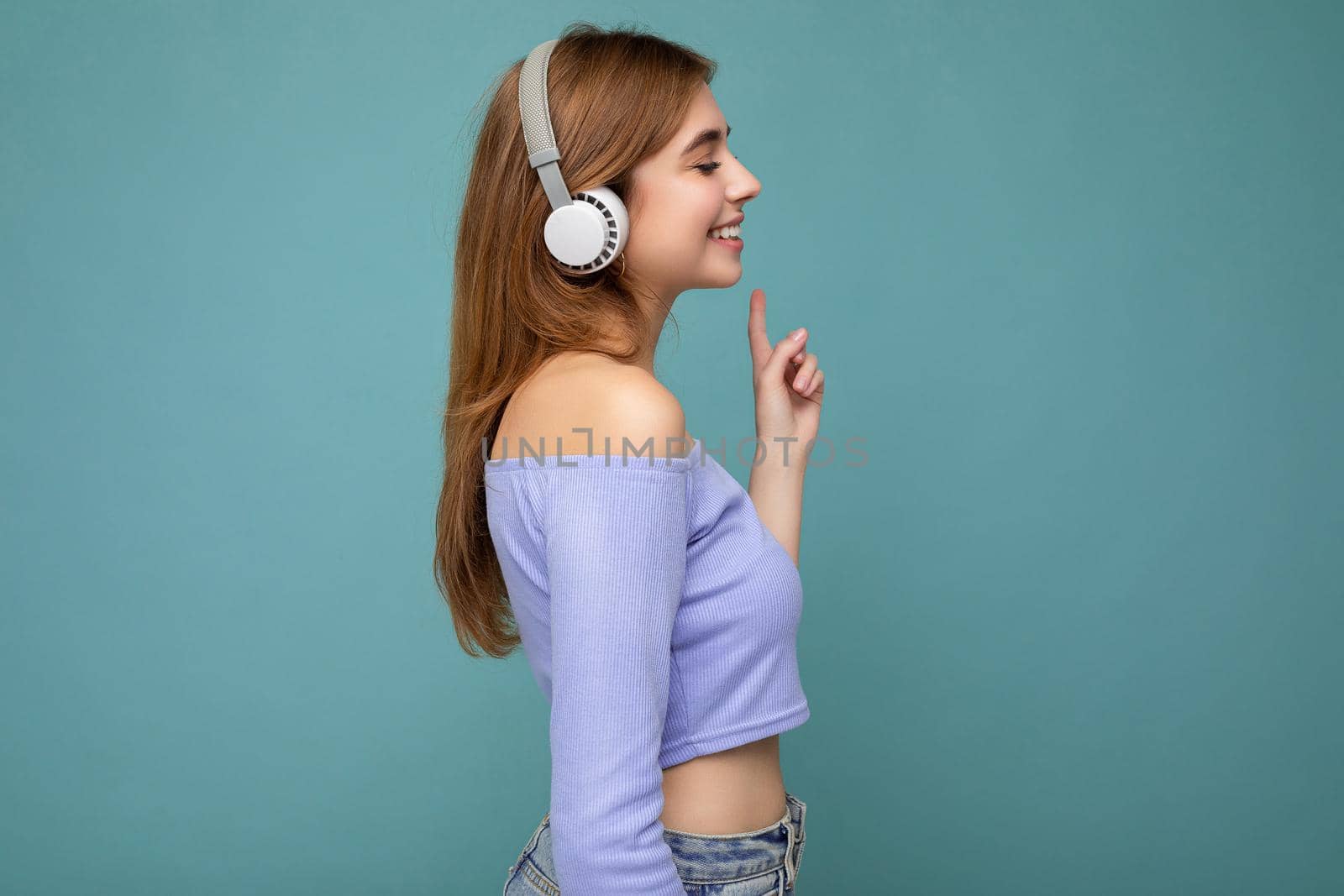 Side-profile photo of beautiful positive smiling young blonde woman wearing blue crop top isolated over blue background wall wearing white wireless bluetooth earphones listening to cool music and looking to the side by TRMK