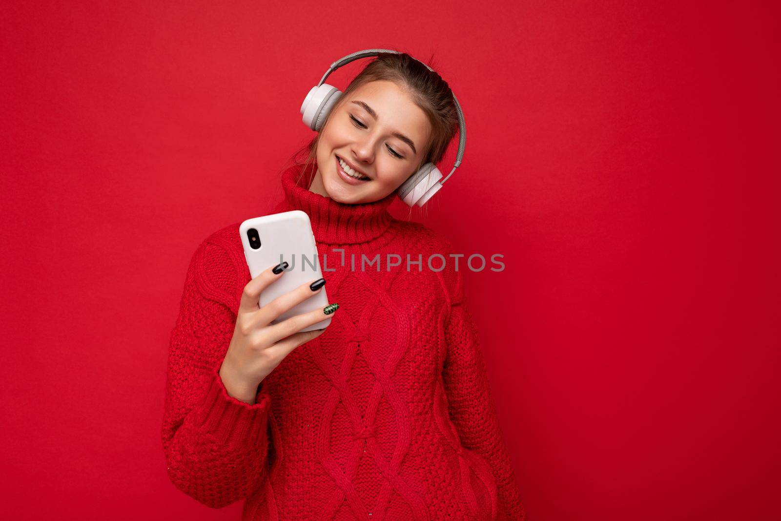 Photo shot of beautiful joyful smiling young female person wearing stylish casual outfit isolated over colorful background wall wearing white bluetooth wireless earphones and listening to music and using mobile phone looking at gadjet display.