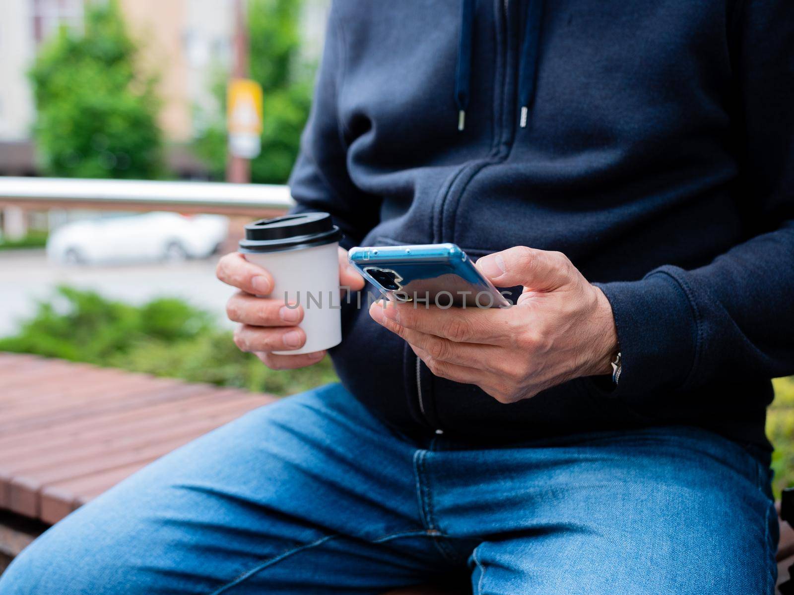 A man sits on a bench drinking coffee from a paper Cup and use a smartphone, close-up. by Utlanov