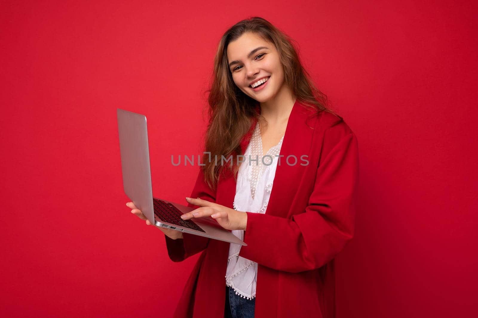 side profile photo of charming smiling confident pretty young lady holding laptop typing text on keyboard wearing red cardigan and white t-shirt isolated on red wall background.