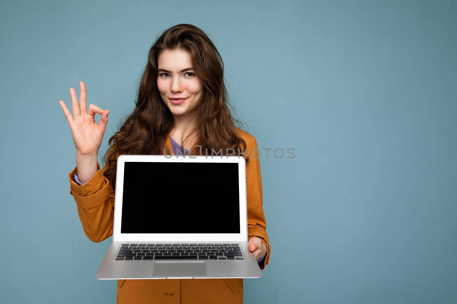 Photo of Beautiful satisfied happy young woman holding computer laptop looking at camera wearing yellow jacket showing ok gesture isolated over blue wall background. mock up, cutout