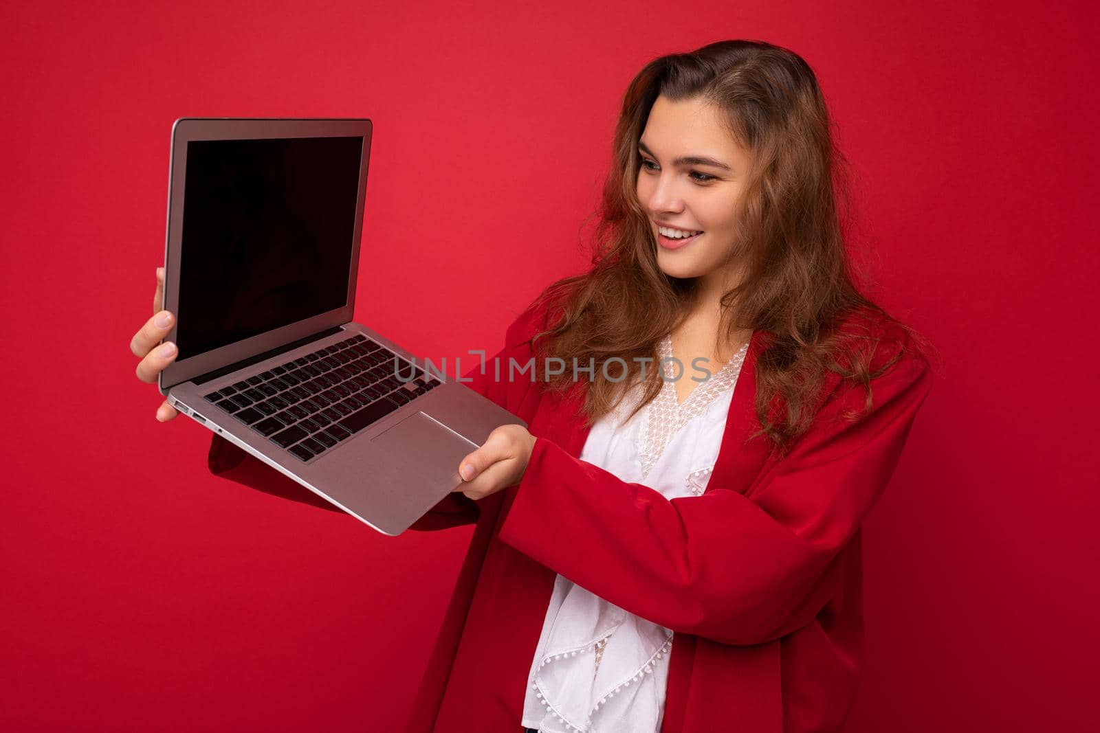 Photo of Beautiful smiling happy young brunette woman holding computer laptop with empty monitor screen wearing red cardigan and white blouse looking at netbook isolated over red background.