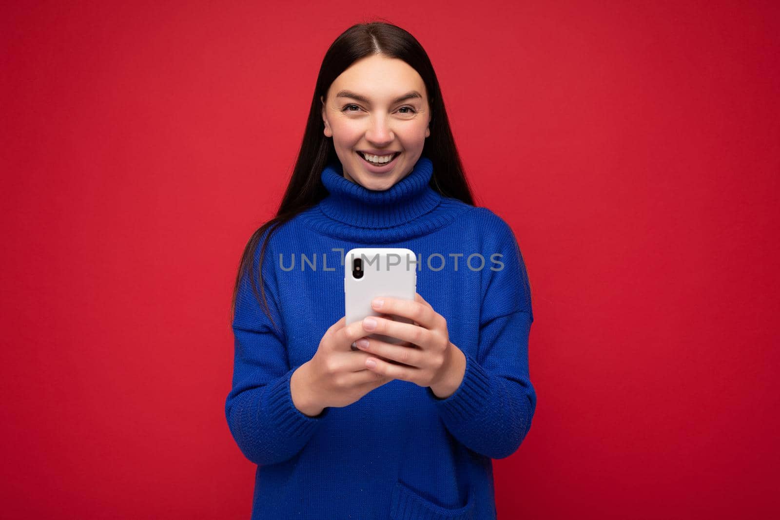 Smiling attractive positive good looking young brunette woman wearing stylish blue warm sweater poising isolated on red background with empty space holding in hand and using mobile phone messaging sms looking at camera.