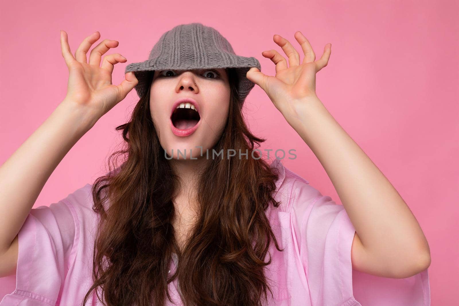 Shot of beautiful emotional young brunette woman standing isolated over pink background wall wearing pink shirt and gray hat and having fun.