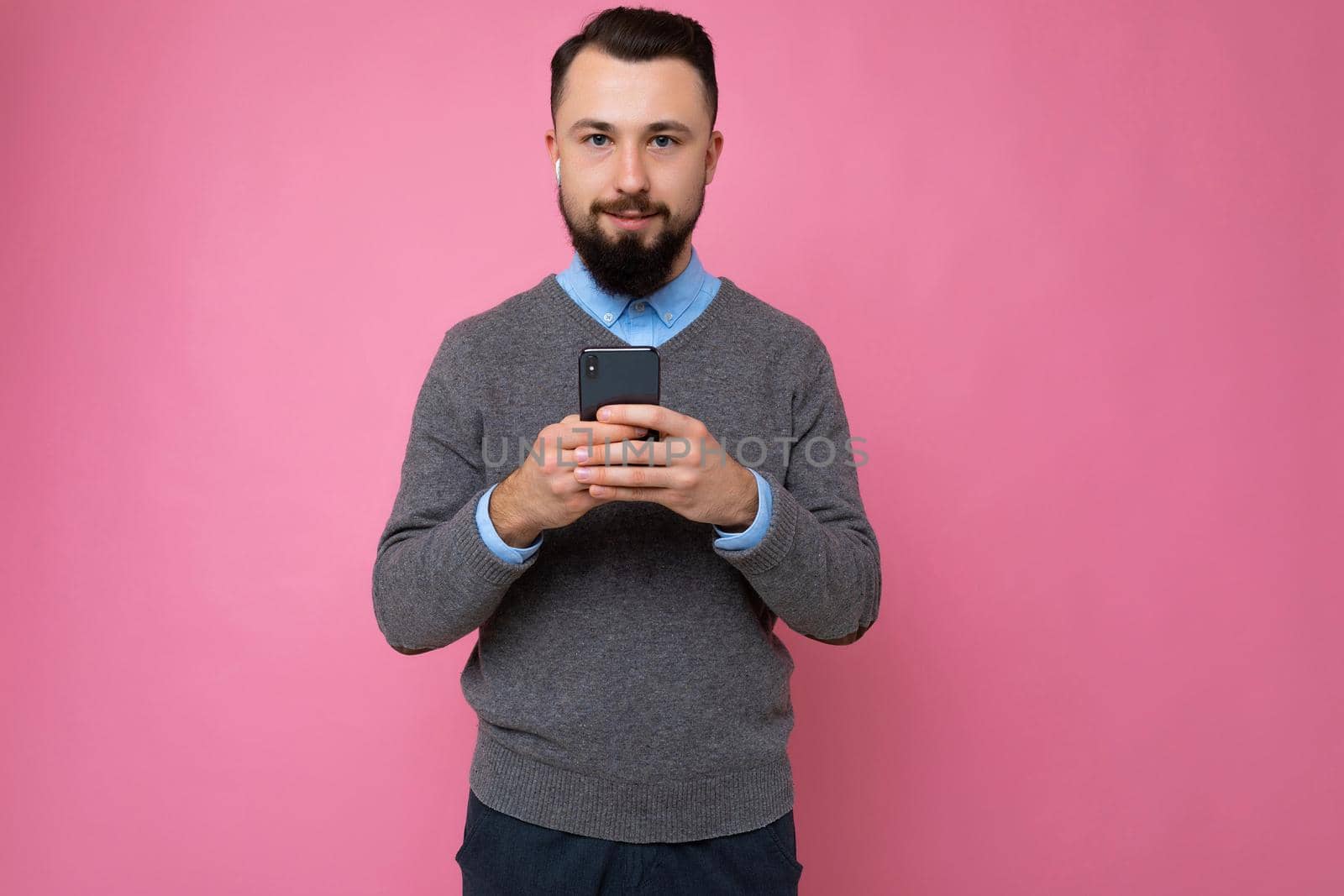 handsome good looking brunet bearded young man wearing grey sweater and blue shirt isolated on pink background with empty space holding in hand and using mobile phone communicating online looking at camera.