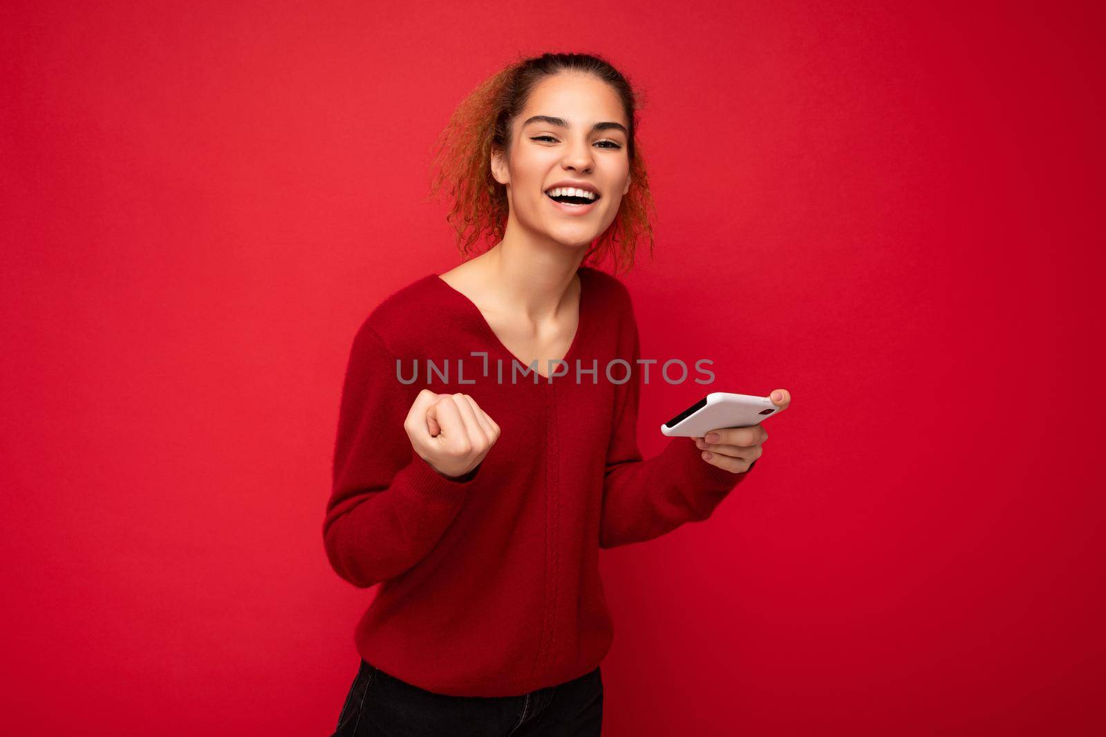 Young emotional woman wearing dark red sweater isolated over red background holding smartphone having fun and showing yes gesture looking at camera.