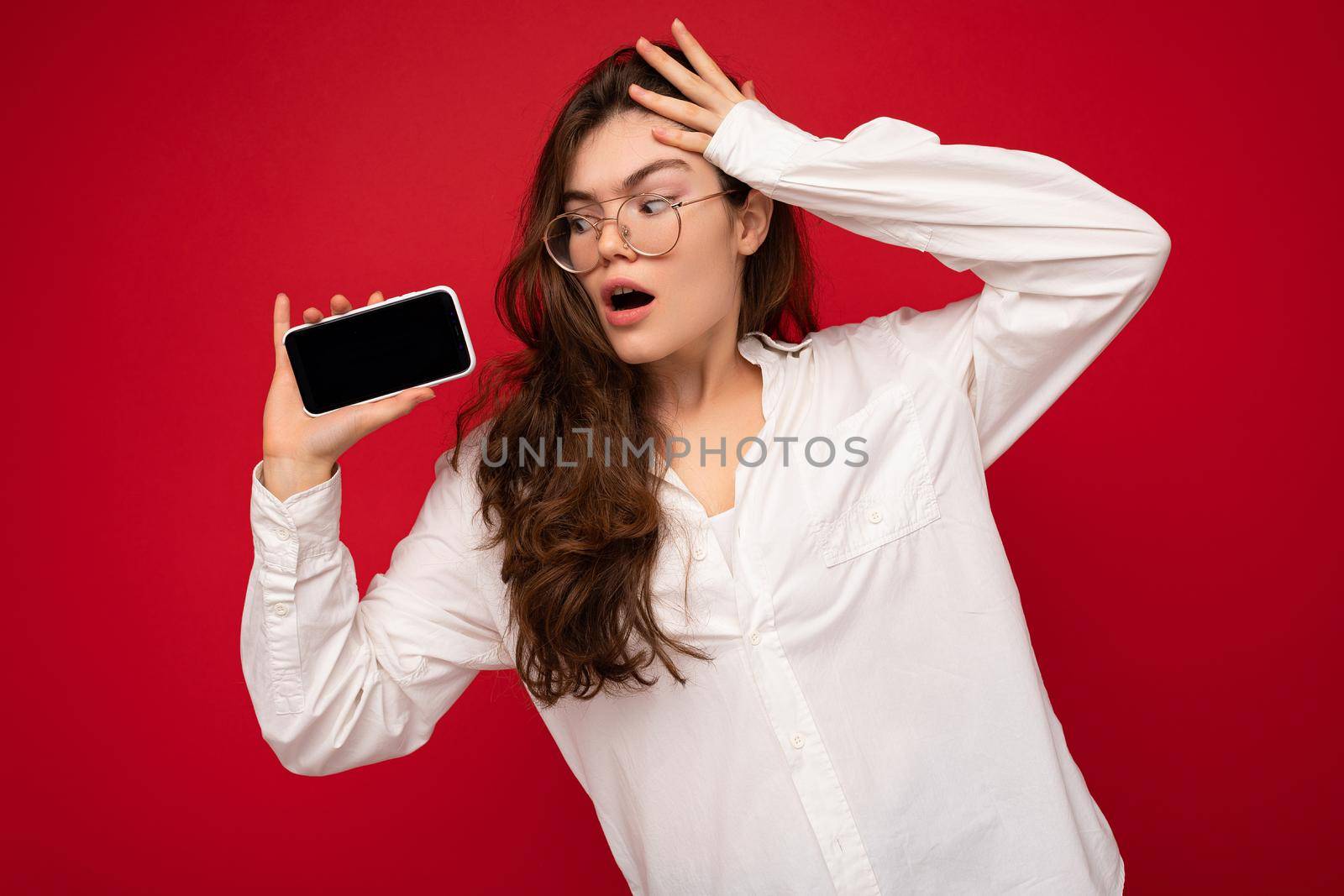 Amazed charming young brunet woman wearing white shirt and optical glasses isolated over red background holding in hand and showing mobile phone with empty screen for mockup looking at gadjet screen.