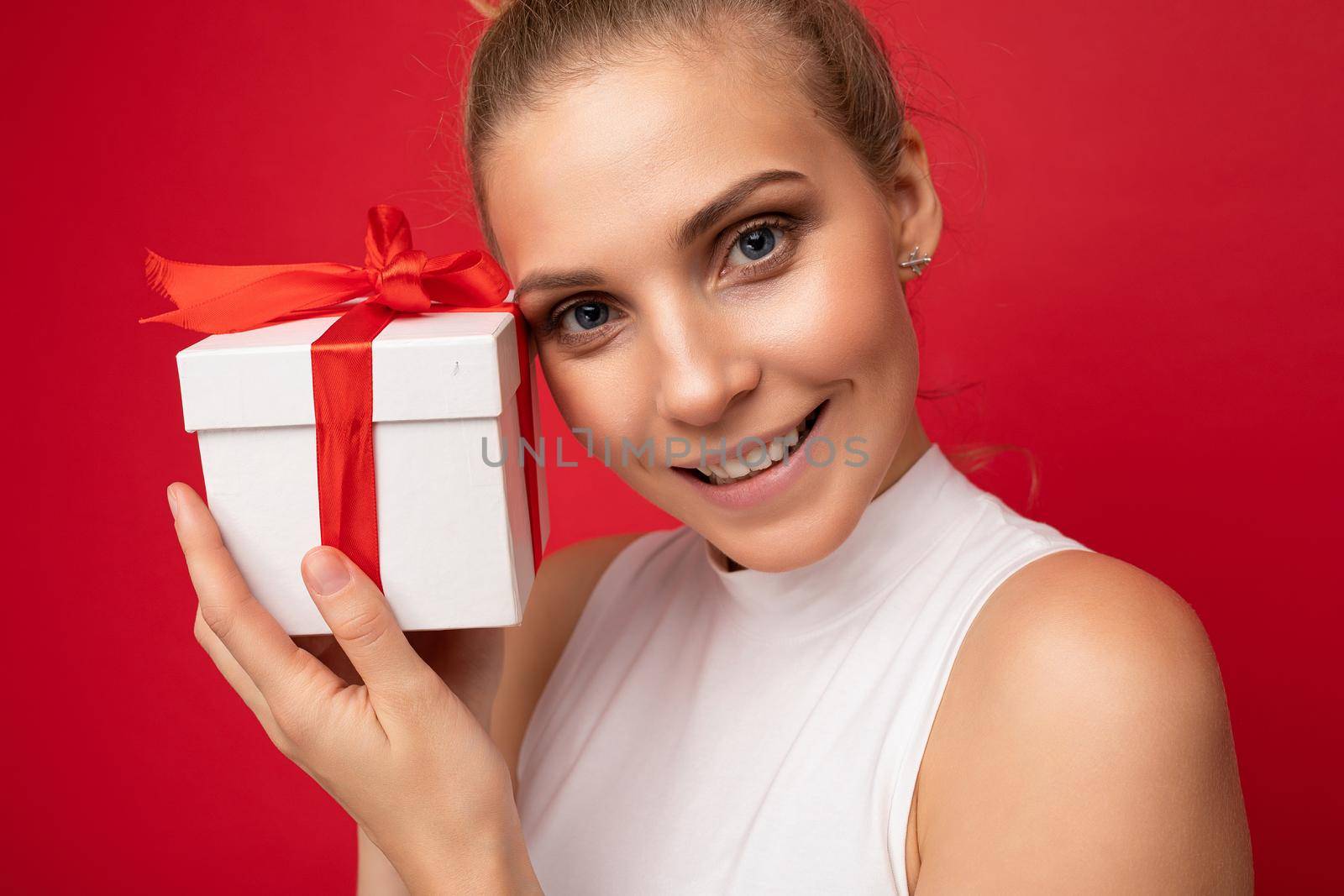Closeup shot of attractive positive smiling young blonde woman isolated over colourful background wall wearing everyday trendy outfit holding gift box and looking at camera.