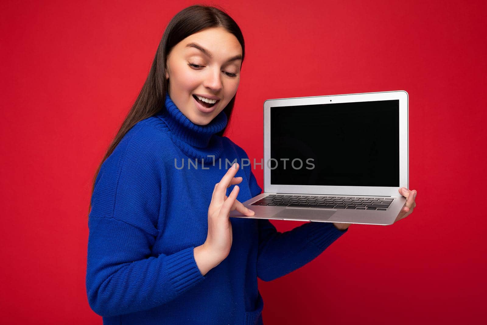 Photo shot of beautiful young brunet woman holding computer laptop looking down in blue sweater isolated over red wall background. Mock up