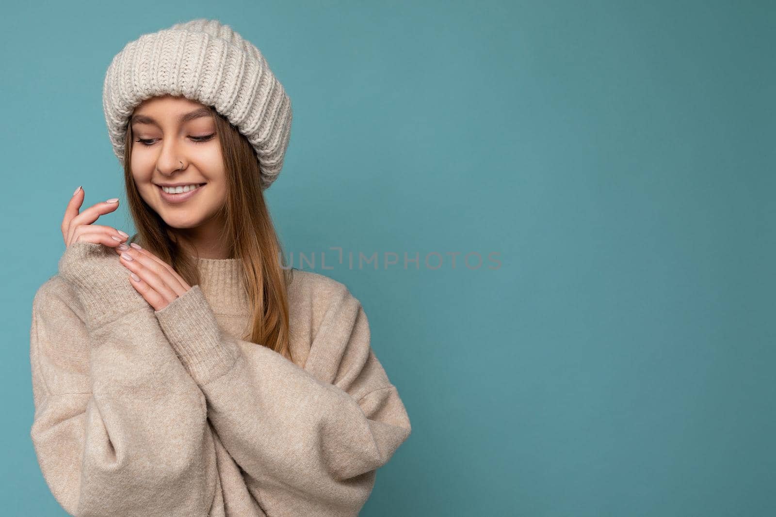 Photo of beautiful happy cute smiling sexy young dark blonde woman isolated over blue background wall wearing beige warm sweater and knitted beige hat looking down. Copy space