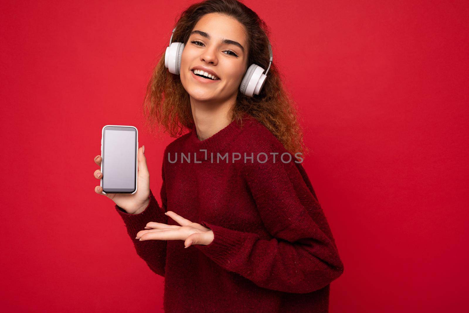 Attractive positive smiling young woman wearing stylish casual outfit isolated on colourful background wall holding and showing mobile phone with empty screen for cutout wearing white bluetooth headphones and having fun looking at camera. copy space