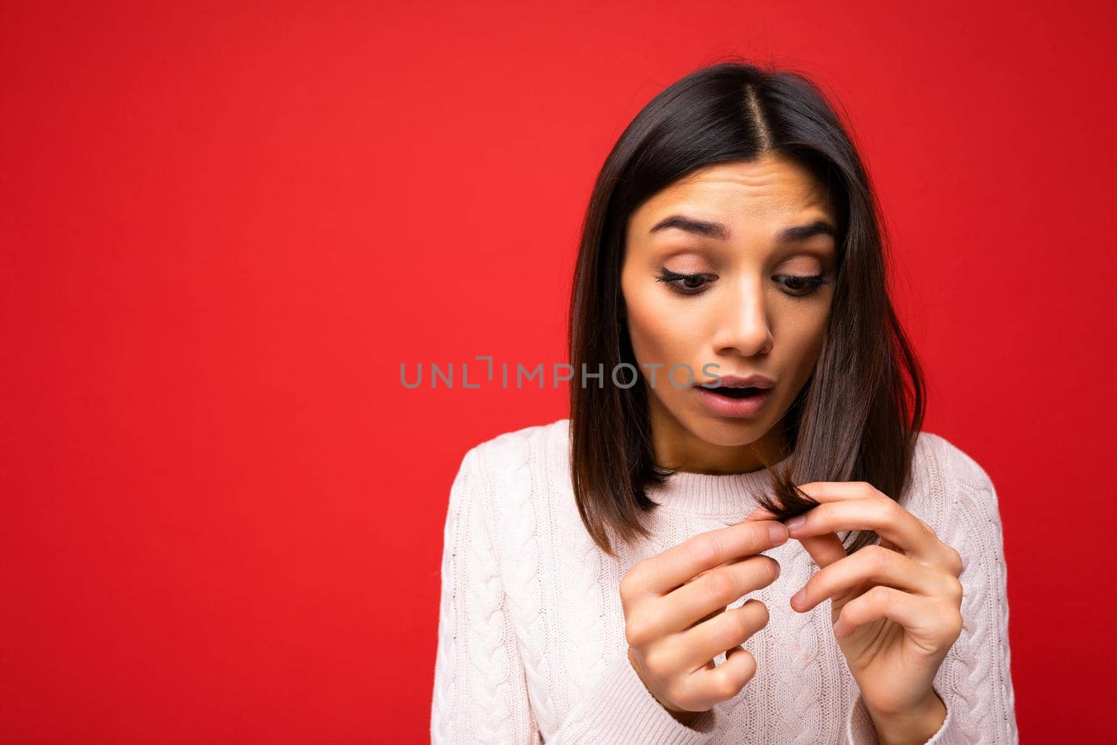 Portrait of beautiful shocked amazed young brunette woman wearing knitted jersey isolated over red background with free space and touching short hair and having split ends.