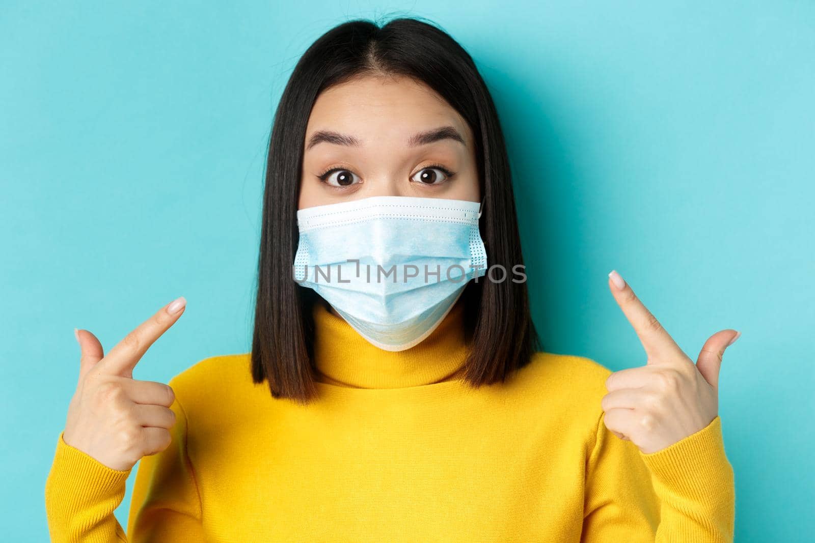 Covid-19, social distancing and pandemic concept. Close up of surprised asian girl pointing at her medical mask and looking amazed at camera, blue background.