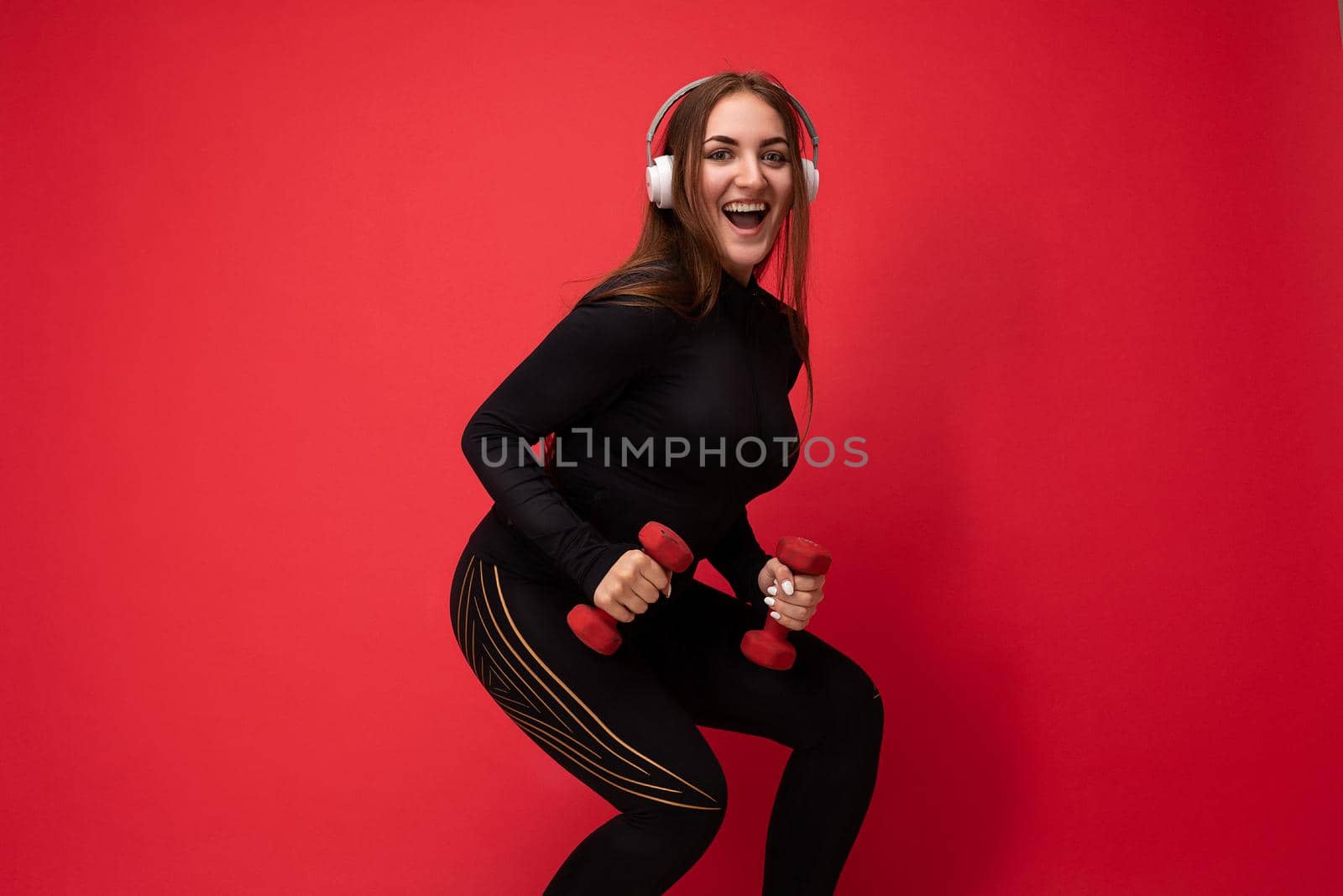 Side-profile photo of beautiful happy smiling young brunet woman wearing black sport clothes isolated on red background wall squating using dumbbells wearing white headsets listening to music looking at camera.