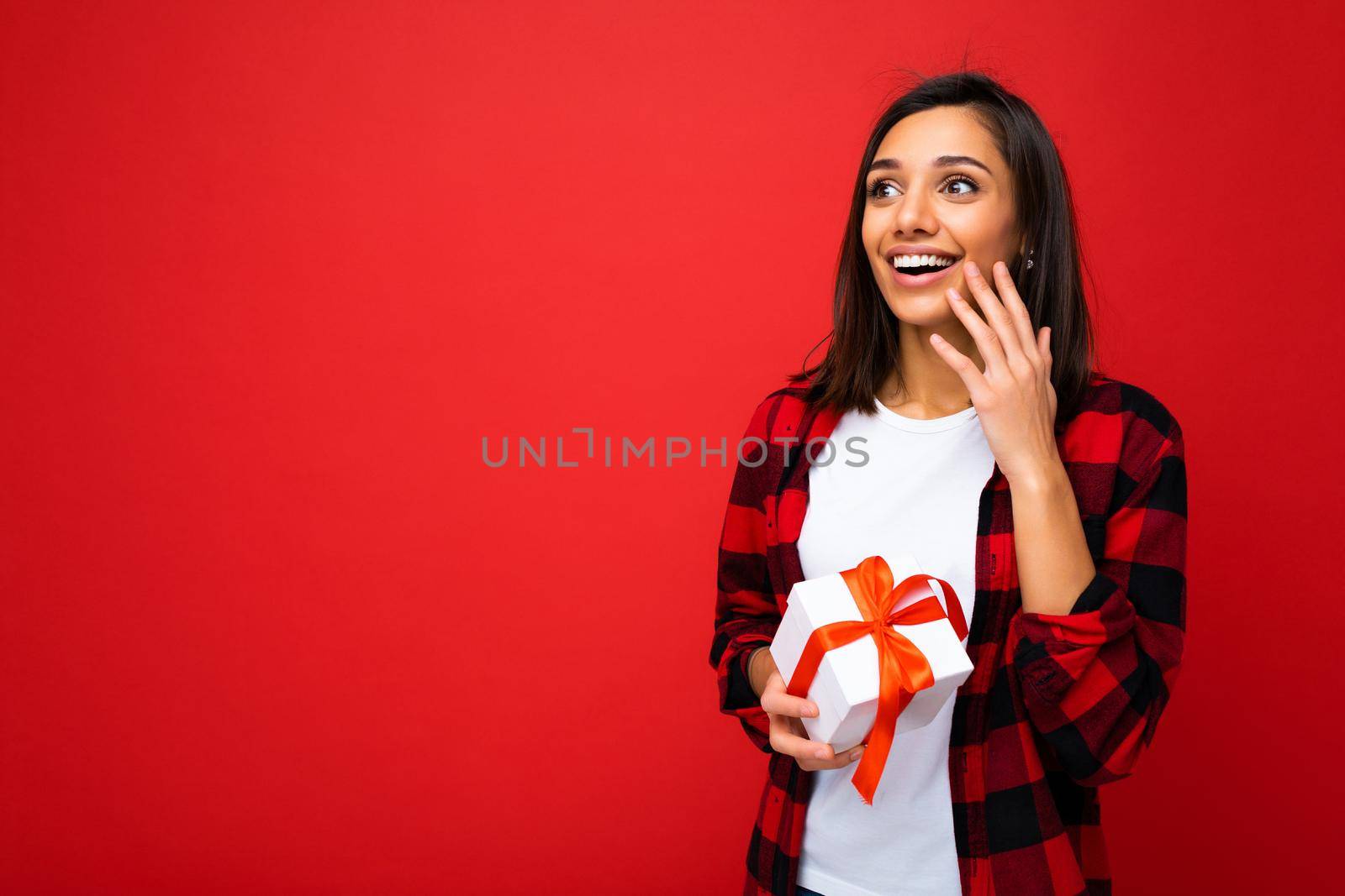 Photo shot of pretty positive surprised young brunet woman isolated over colourful background wall wearing trendy outfit look holding gift box and looking to the side. Free space