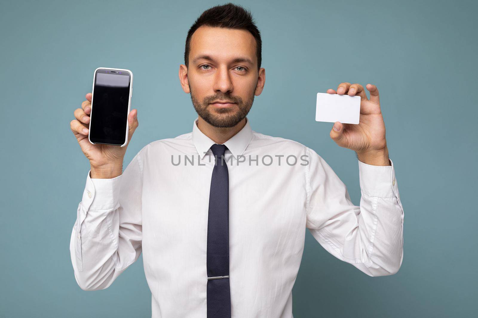 Photo of handsome good looking brunet man with beard wearing casual white shirt and tie isolated on blue background with empty space holding in hand and showing mobile phone with empty screen for mockup and credit card looking at camera.