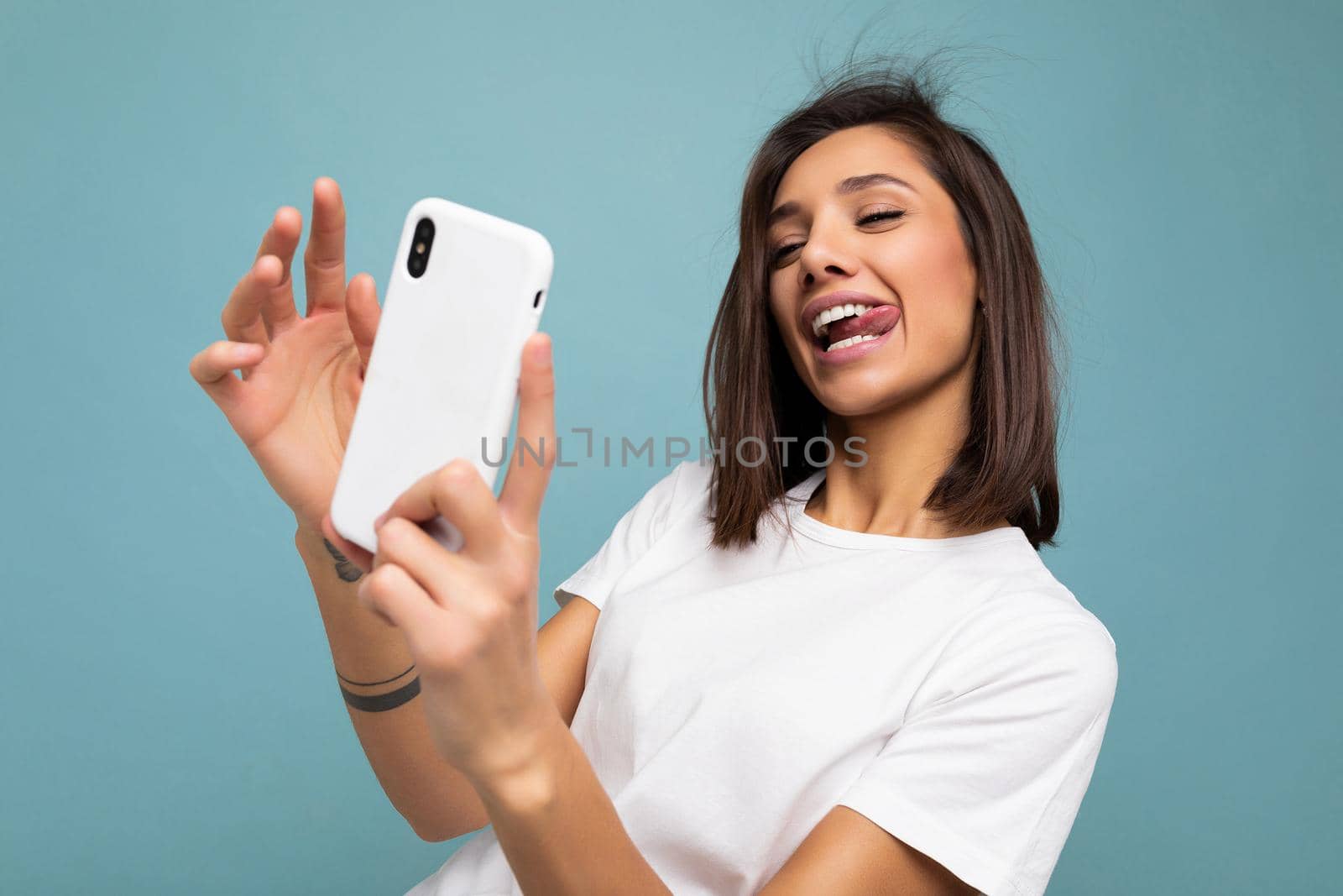 Photo of sexy beautiful smiling happy young brunette woman wearing casual white t-shirt isolated over wall blue background holding and using mobile phone taking selfie looking at gadjet screen and showing tongue.