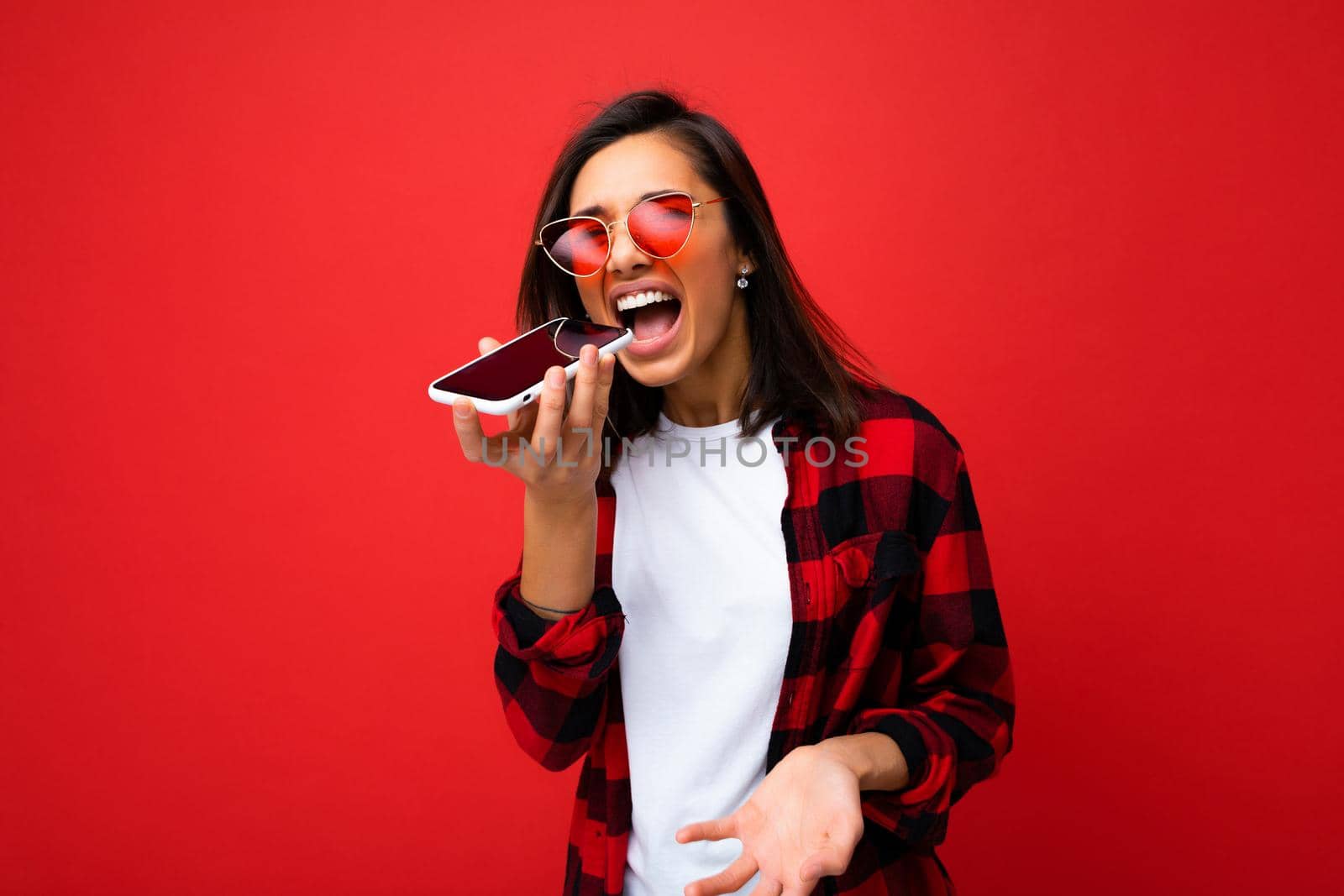 photo of beautiful emotional young brunet woman wearing stylish red shirt white t-shirt and red sunglasses isolated over red background using mobile phone recording voice message and shouting.