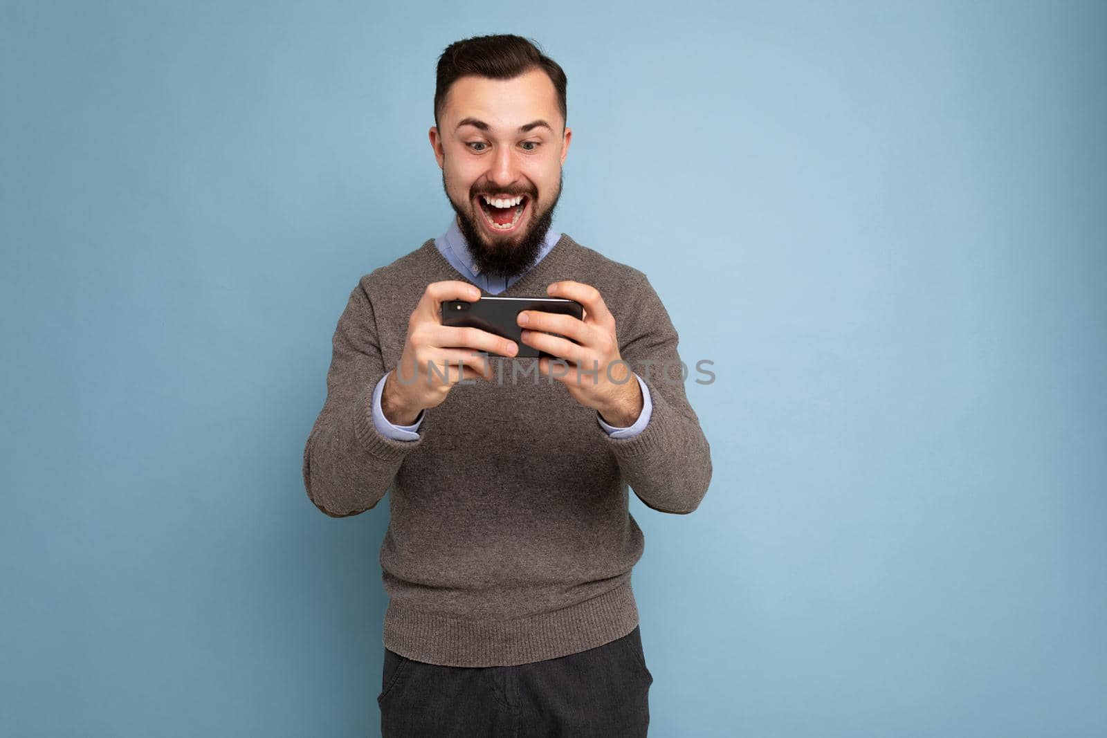 Shot of happy amazing shocked handsome young brunette unshaven man with beard wearing everyday grey sweater and blue shirt isolated on background wall holding smartphone playing games via mobile phone looking at phone screen display and having fun.