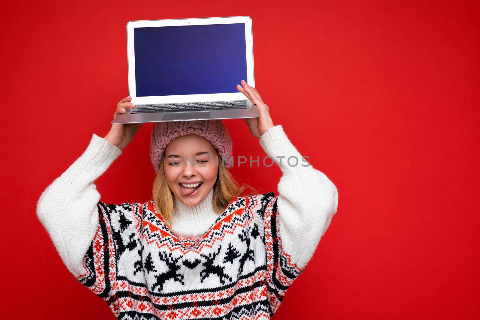 Photo shot of beautiful smiling blonde young woman holding computer laptop with empty monitor screen with mock up and copy space wearing knitted winter hat and sweater showing tongue isolated over red wall background.
