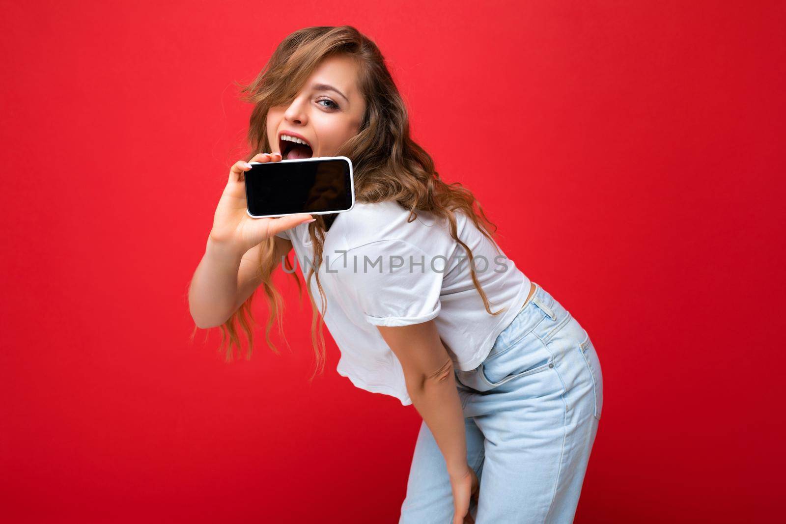 Photo of beautiful smiling young blonde woman good looking wearing white t-shirt standing isolated on red background with copy space holding phone showing smartphone in hand with empty screen display for mockup looking at camera.