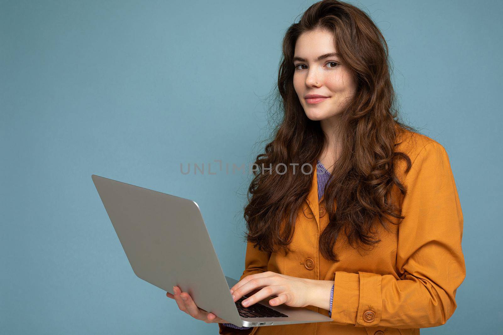 Photo of beautiful young woman holding computer laptop looking at camera isolated over colourful background.