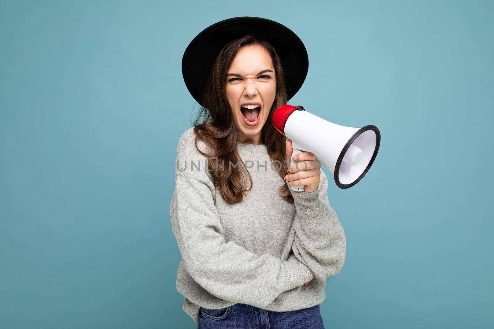 Photo of young beautiful positive brunette woman with sincere emotions wearing stylish black hat and grey sweater isolated over blue background with copy space and screaming in magaphone.