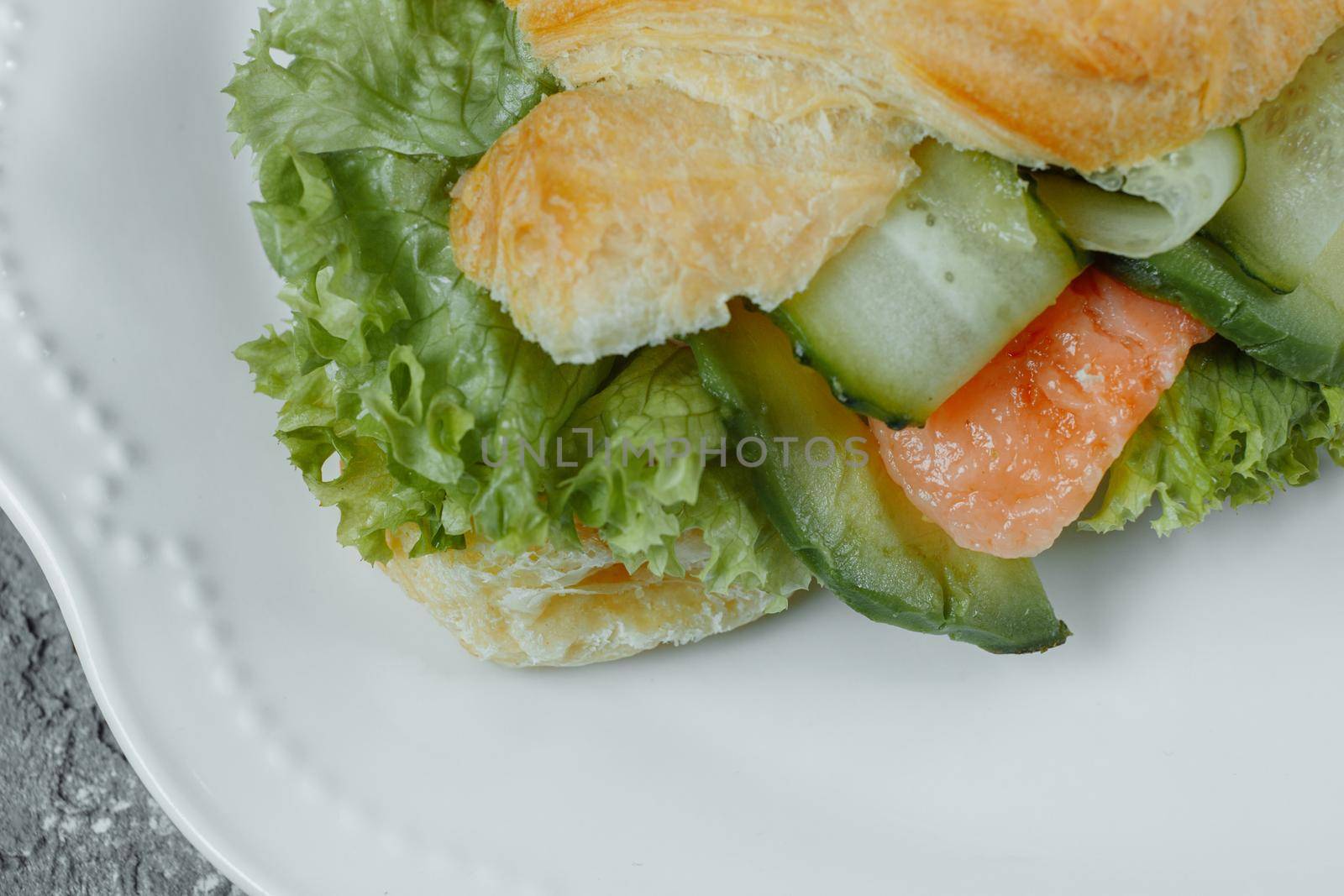 Croissant sandwich with red fish, avocado, fresh vegetables and arugula on black shale board over black stone background. Healthy food concept.