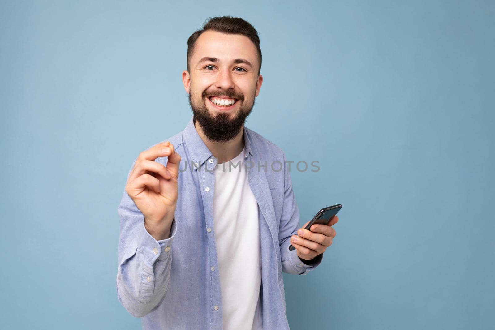 Closeup portrait of positive emotional good looking young brunet bearded man wearing casual blue shirt and white t-shirt poising isolated on blue background with empty space holding in hand mobile phone looking at camera and explaining smth.