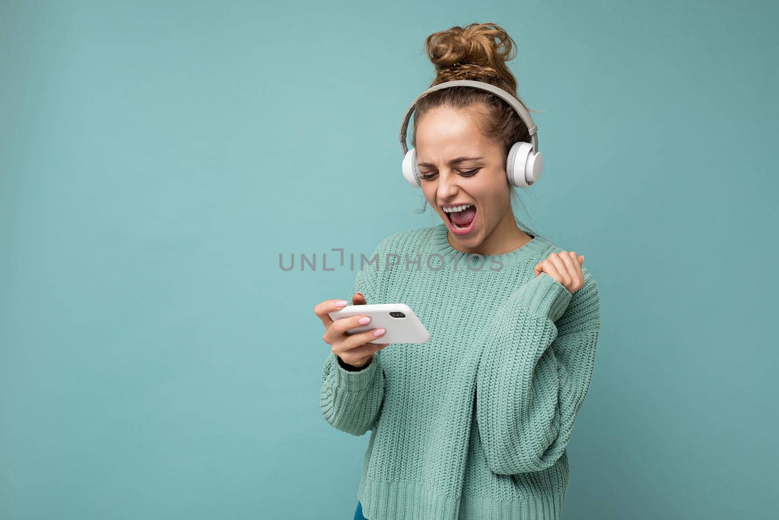 Attractive emotional positive young woman wearing blue sweater isolated over blue background wearing white bluetooth wireless headphones and listening to music and using mobile phone playing online games looking at gadjet screen and having fun. copy space