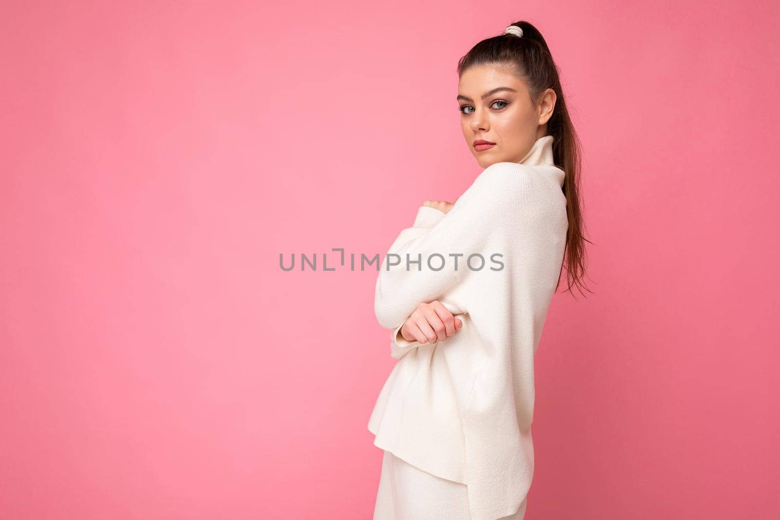 Side-profile shot of young attractive shy stylish brunette woman wearing casual white sweater poising isolated on pink background wall. Copy space, free space, empty space