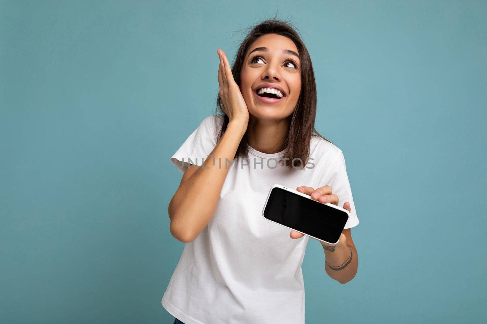 Photo of pretty positive amazed young brunette woman good looking wearing white t-shirt standing isolated on blue background with copy space holding smartphone showing phone in hand with empty screen screen for cutout looking up.