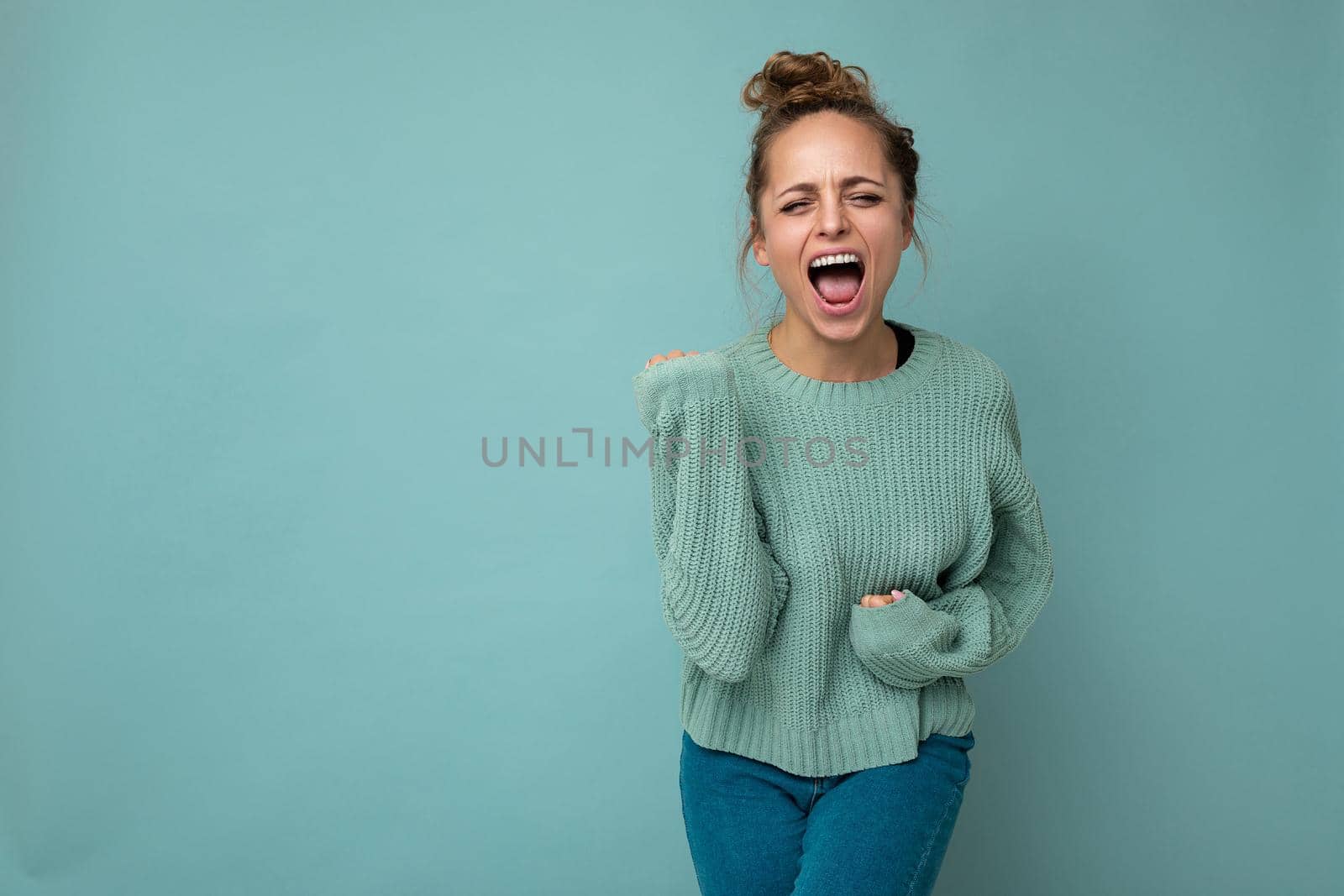 Shot of young funny joyful happy pretty blonde woman wearing blue jersey isolated on blue background with empty space and celebrating victory.