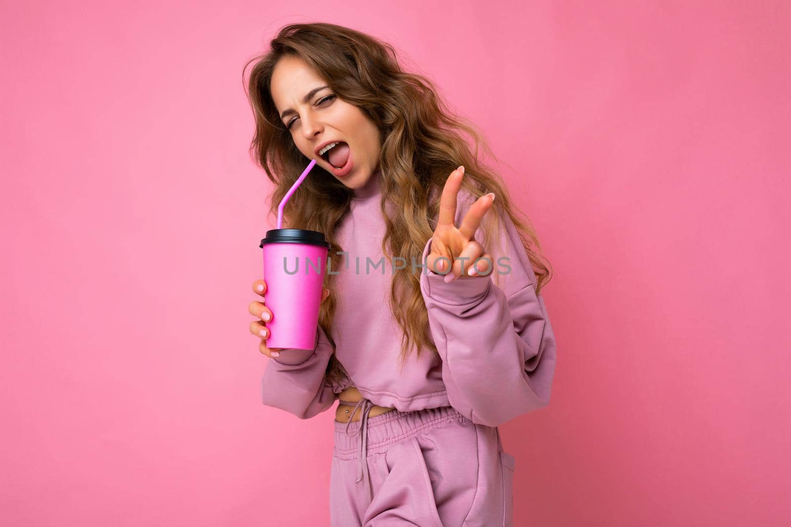 Charming young happy smiling blonde woman wearing stylish clothes isolated over colourful background wall holding paper cup for mockup drinking milkshake looking at camera and showing peace gesture.