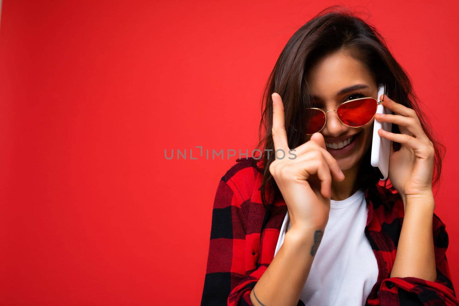 Closeup photo of sexy pretty happy young brunet woman wearing stylish red shirt white t-shirt and red sunglasses isolated over red background communicating on mobile phone looking at camera.
