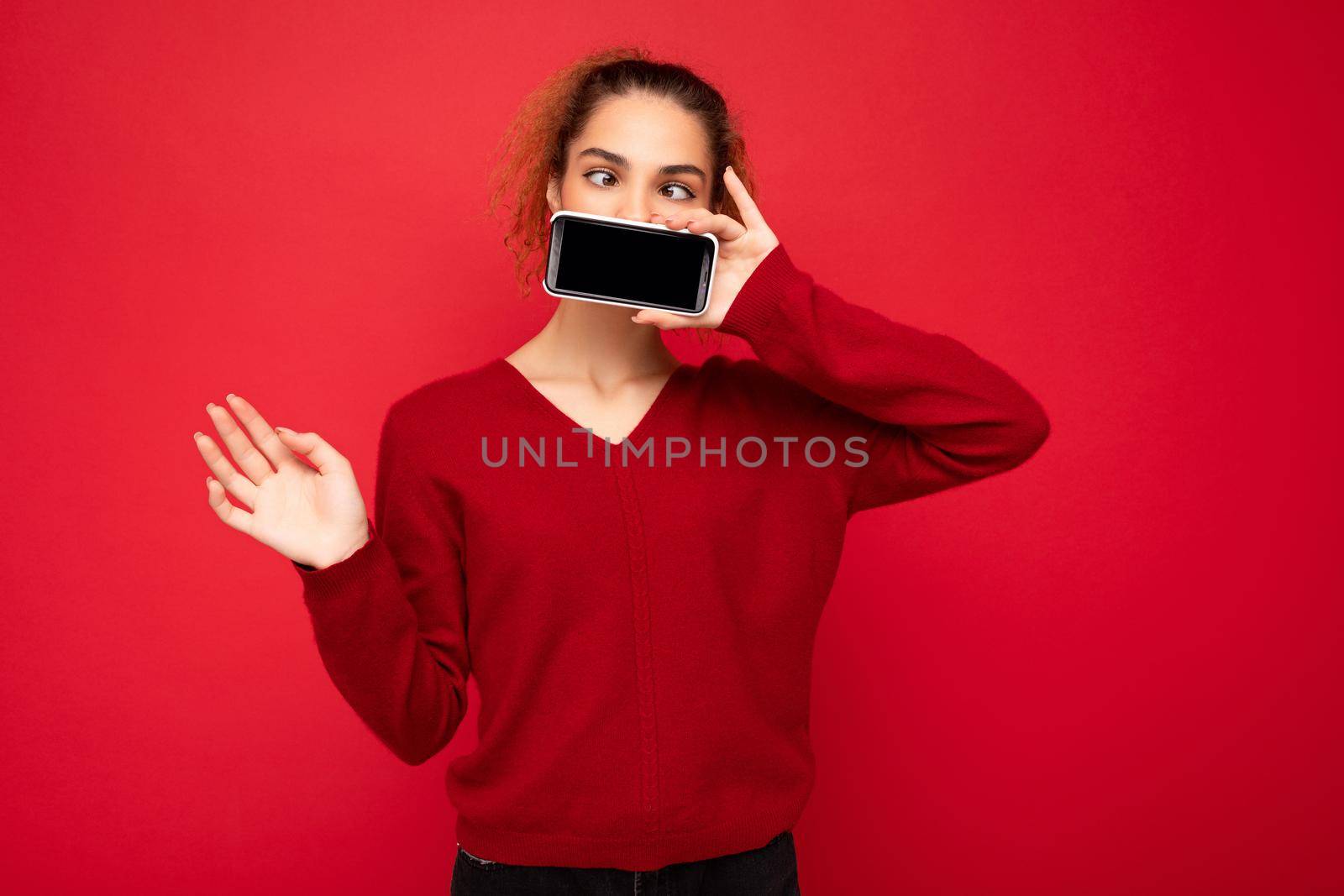 Photo of happy funny young female person wearing dark red sweater isolated over red background holding smartphone and showing mobile phone screen with copy space for cutout.
