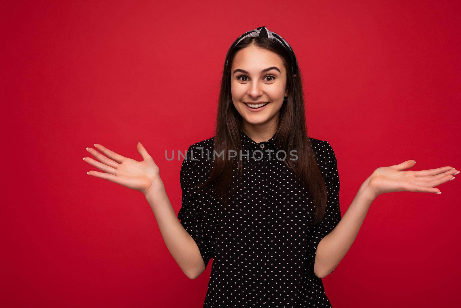 Photo of beautiful smiling positive brunette girl standing isolated over red wall wearing casual stylish black clothes looking at camera and showing two open palms.