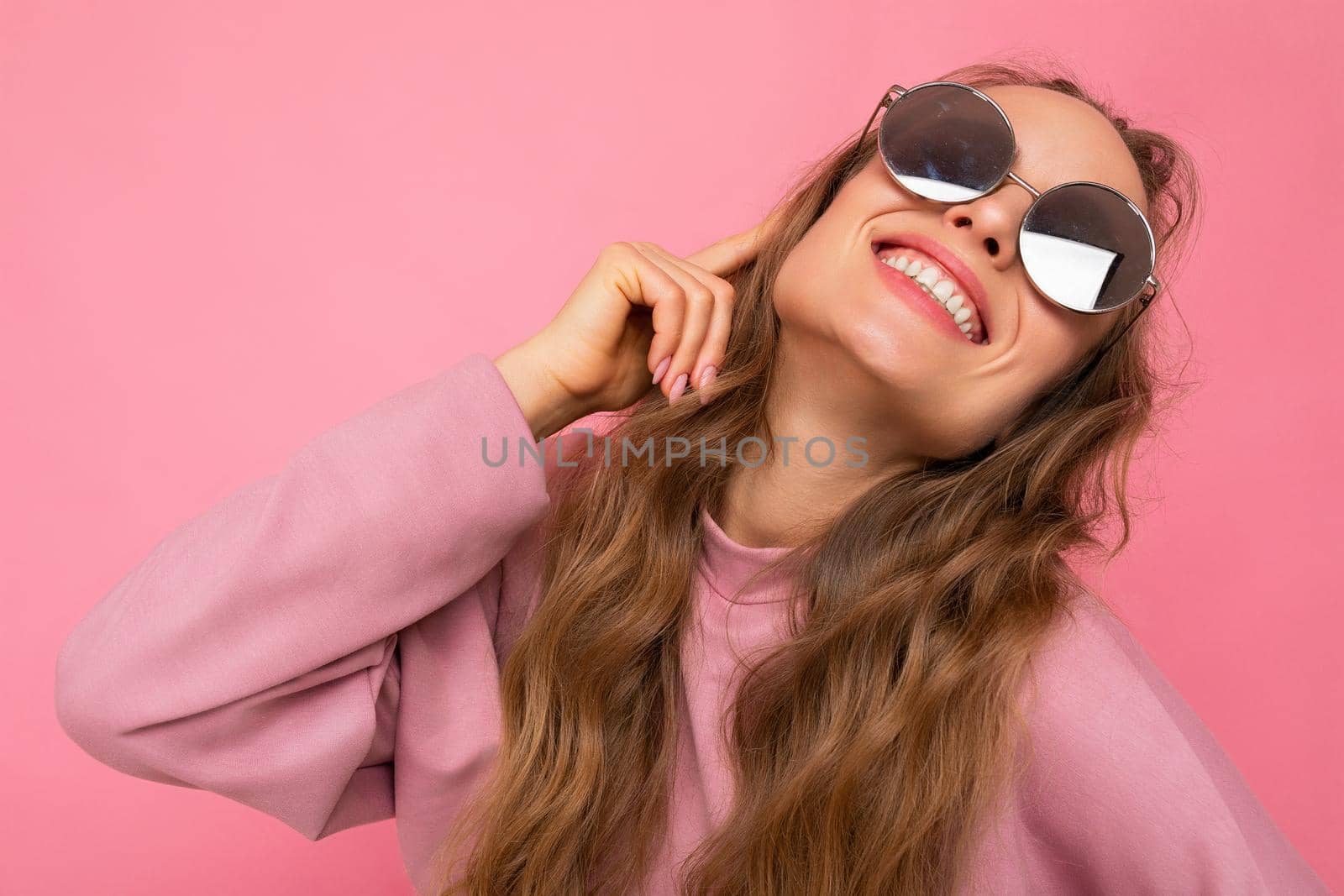 Shot of beautiful positive smiling happy young dark blonde curly woman isolated over pink background wall wearing casual pink sport clothes and stylish sunglasses looking up.