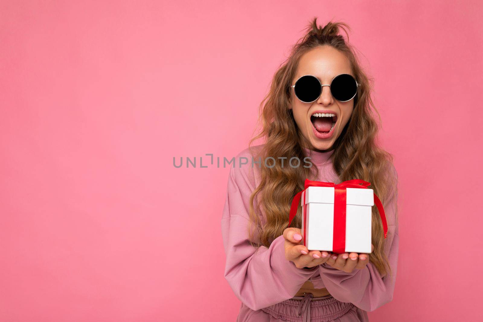 Pretty amazed shocked happy young blonde curly woman isolated over pink background wall wearing pink sport clothes and sunglasses holding gift box looking at camera. Empty space