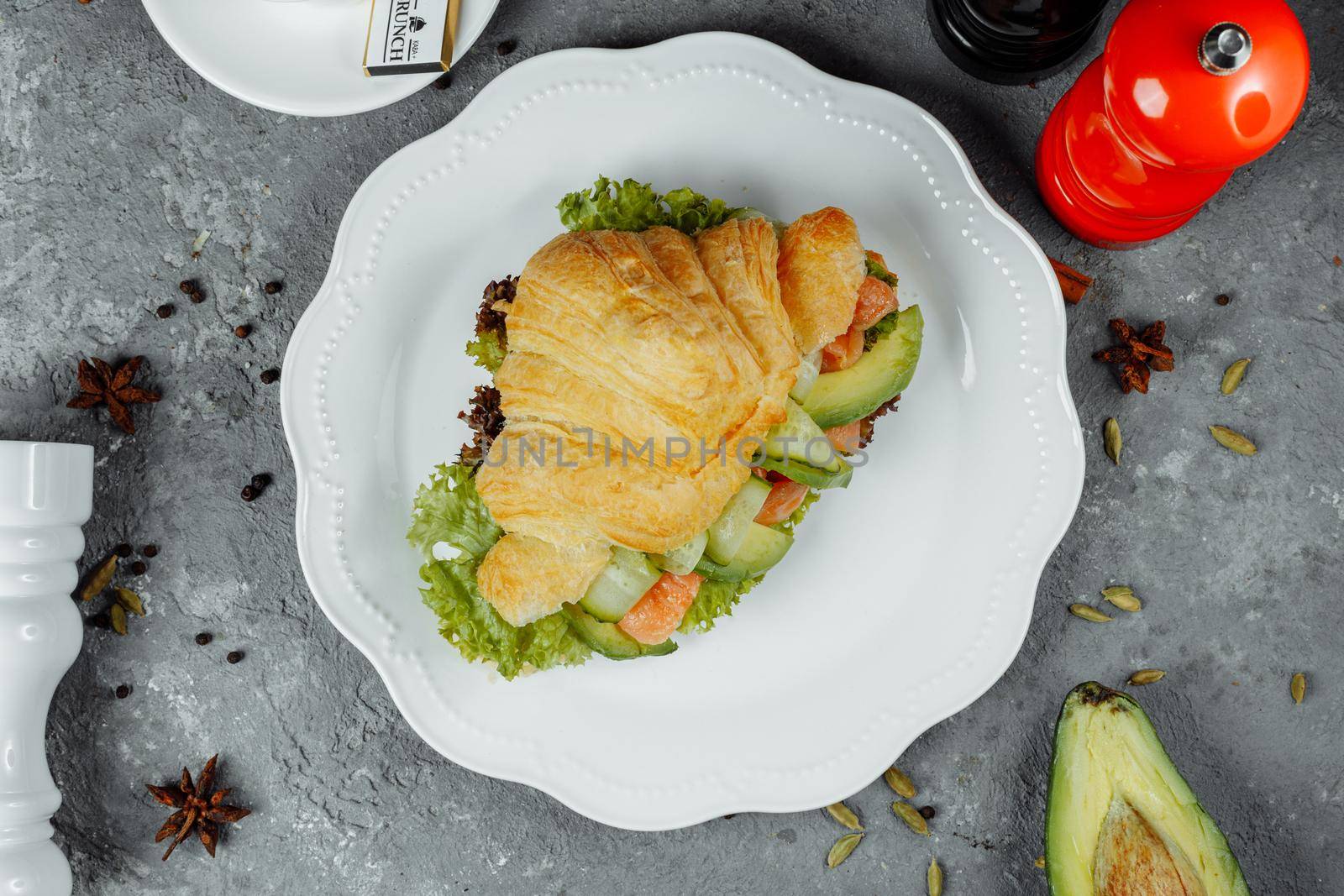 Croissant sandwich with red fish, avocado, fresh vegetables and arugula on black shale board over black stone background. Healthy food concept.