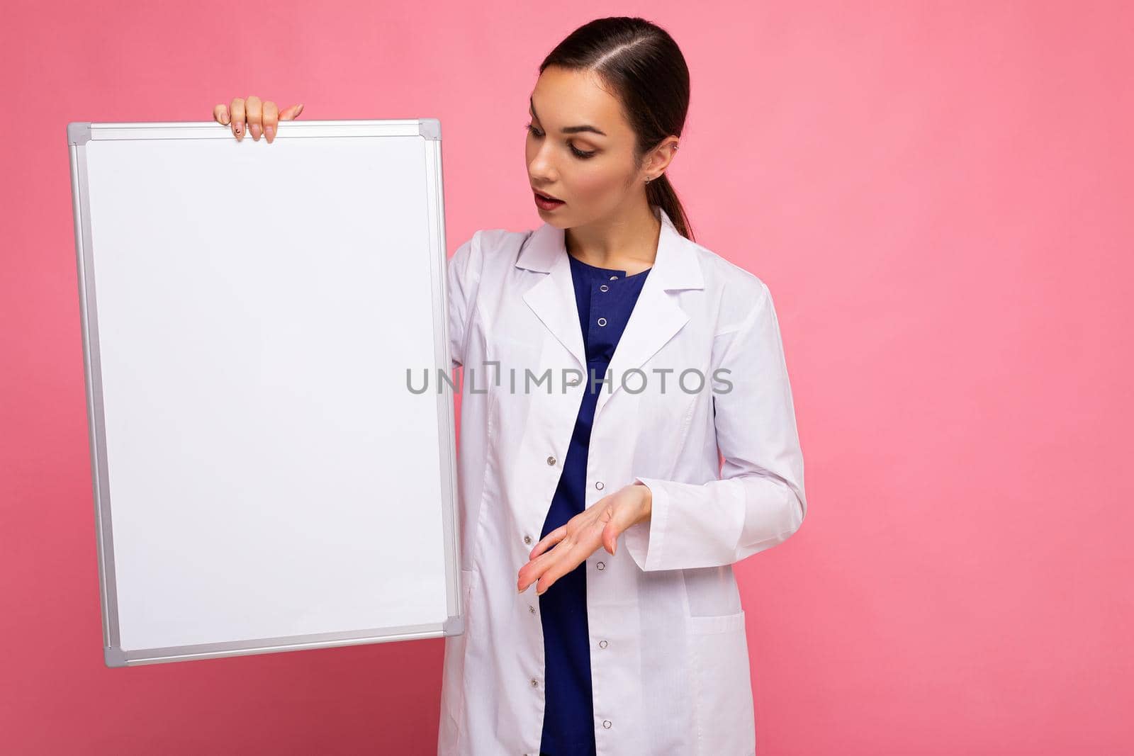 Young of beautiful happy brunet woman wearing medical white coat holding white magnetic board for mockup isolated on pink background with empty space.