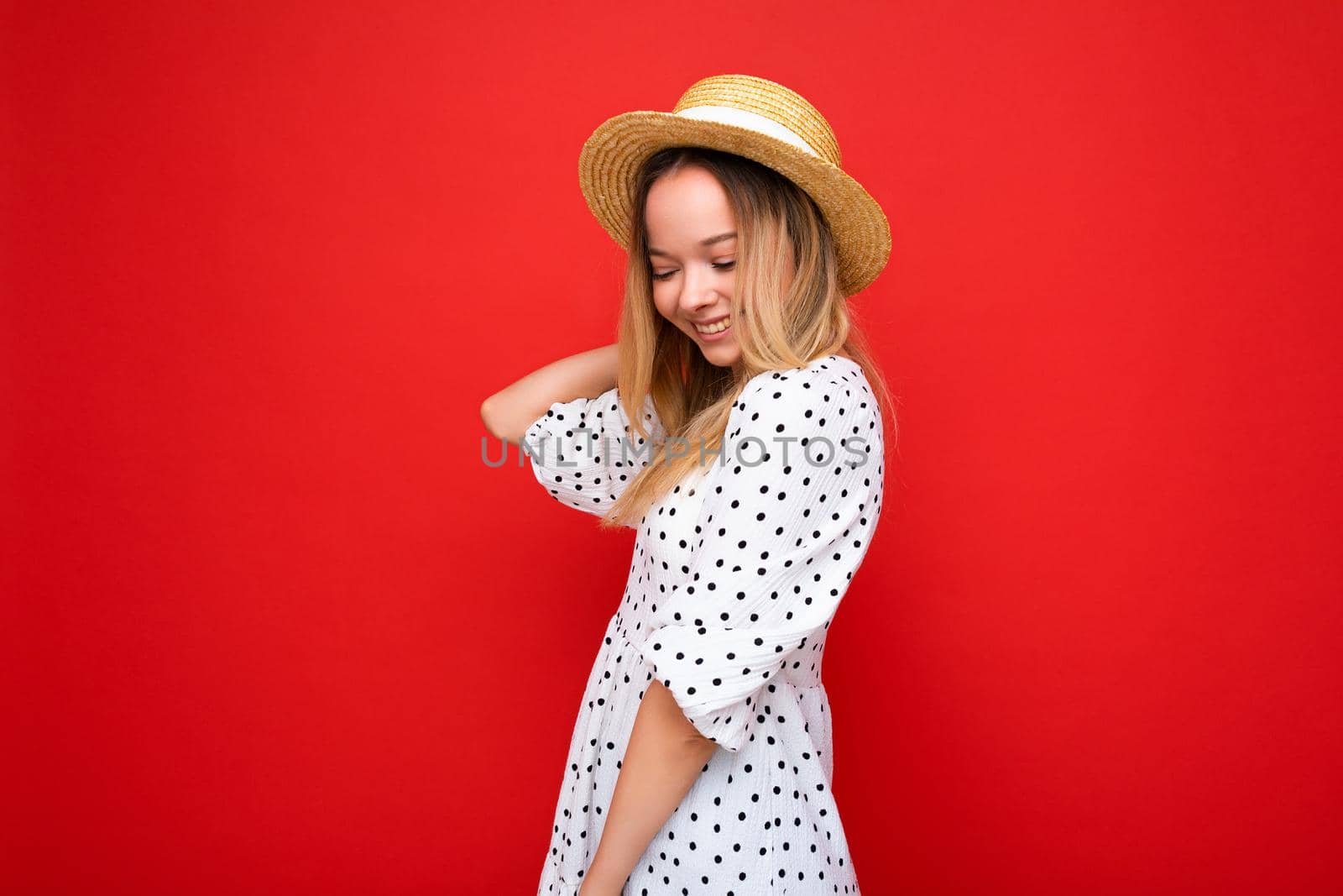 Portrait of young beautiful smiling hipster blonde woman in trendy summer dress and straw hat. Sexy carefree female person posing isolated near red wall in studio. Positive model with natural makeup. Copy space by TRMK