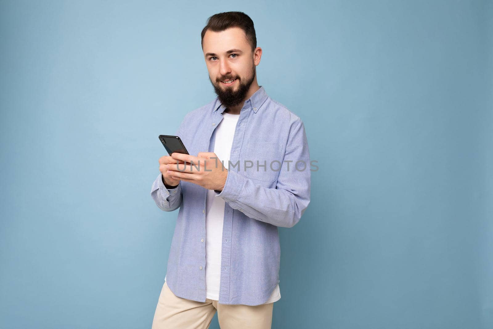 handsome young brunette unshaven man with beard wearing stylish white t-shirt and blue shirt isolated over blue background with empty space holding in hand and using phone messaging sms looking at camera.
