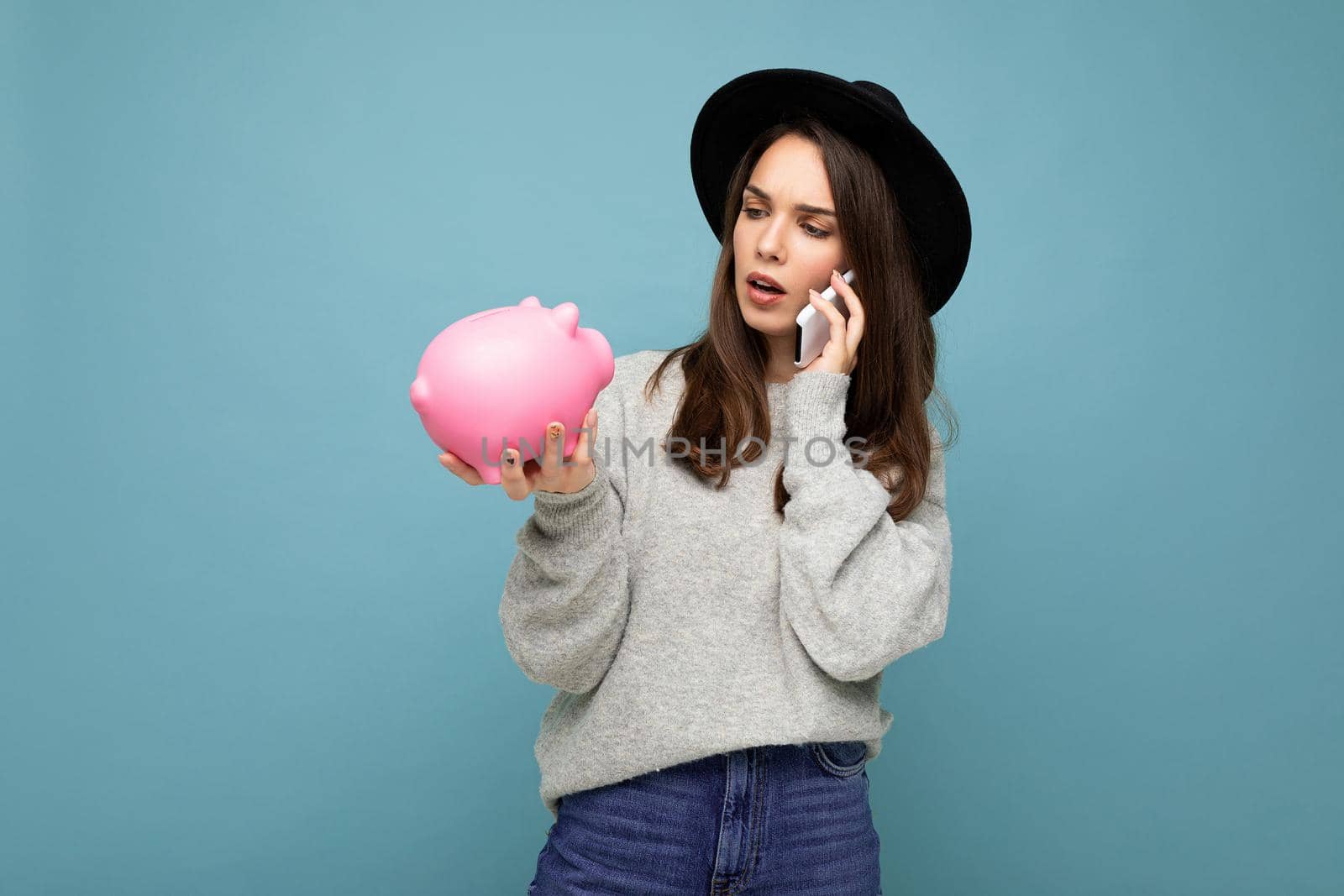 Photo of asking serious thoughtful young attractive pretty woman with dark streight long hair wearing casual grey sweater and black hat isolated over blue background with free space, holds pink pig savings box and speaks on smartphone online.