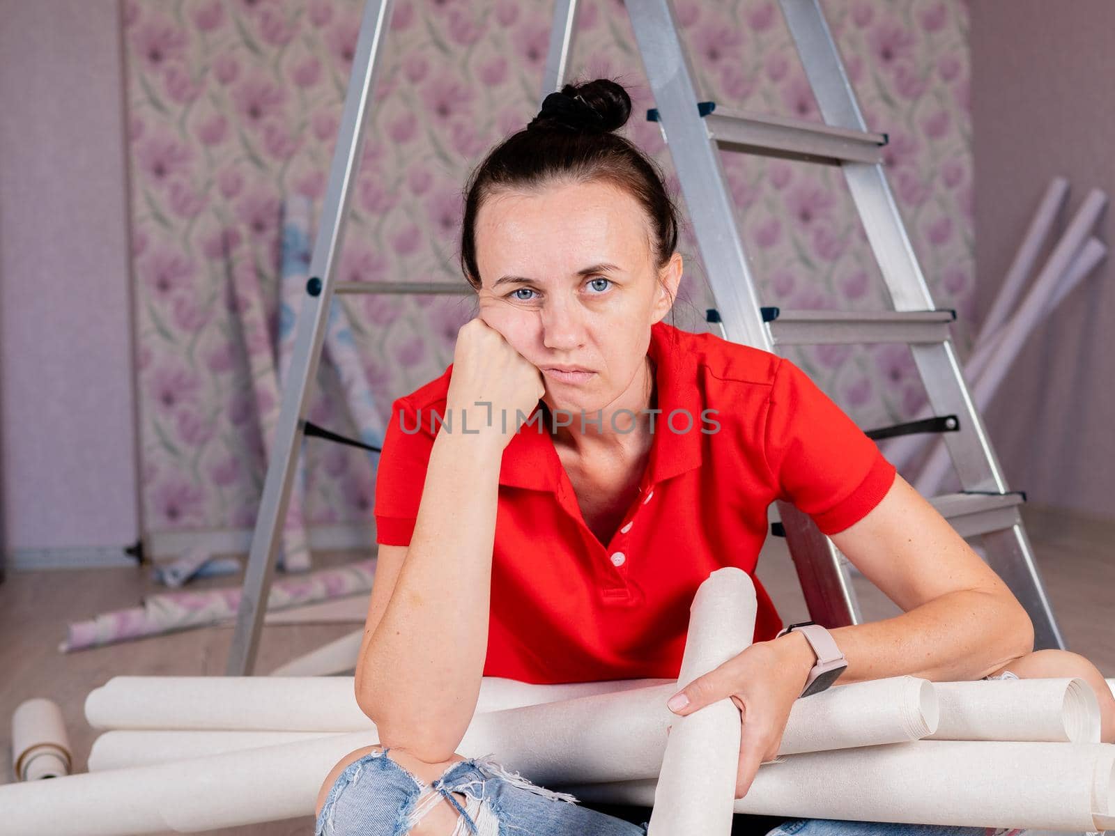 A woman sits on the floor and is sad because of the heavy repairs in the apartment. Wallpapering in the room. by Utlanov
