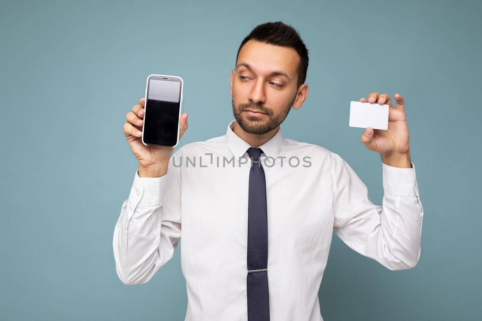 Photo of handsome good looking brunet man with beard wearing casual white shirt and tie isolated on blue background with empty space holding in hand and showing mobile phone with empty screen for mockup and credit card looking to the side.