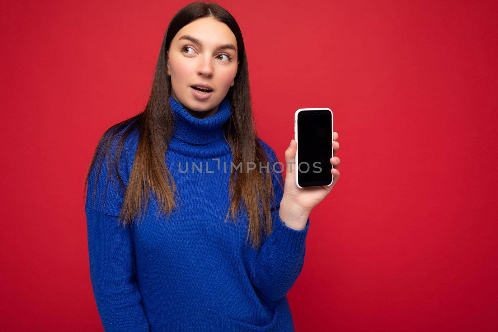 beautiful happy young brunette woman wearing casual blue sweater isolated over red background with empty space holding in hand mobile phone and showing smartphone with empty screen for mockup looking to the side.